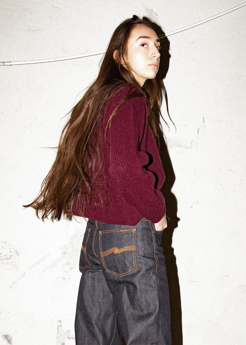 A woman with long dark brown hair is shown from the side, wearing a dark burgundy ribbed knit sweater and dark gray wide-leg jeans, standing against a textured off-white wall.