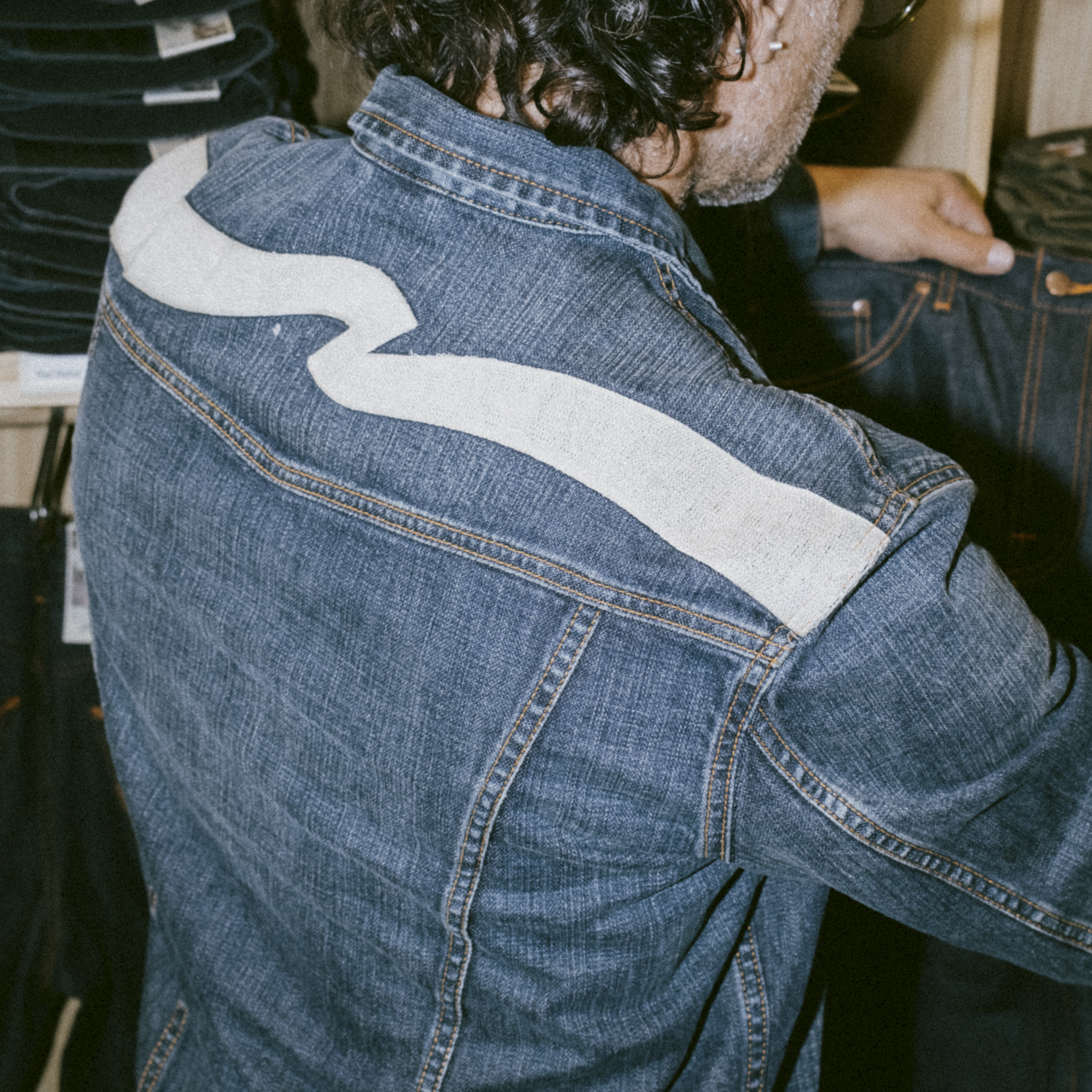 Man photographed from the back wearing a faded blue denim jacket with the signature Nudie Jeans Ecru Waves design across the back shoulders.
