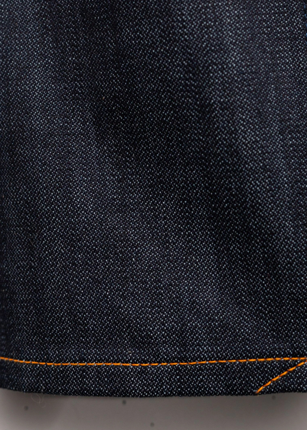 Close-up of dark indigo denim fabric with orange contrast stitching along the hem.