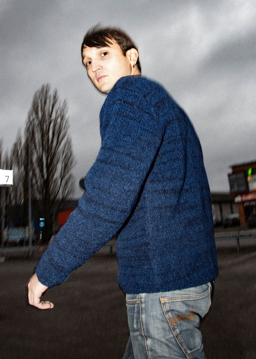 A man wearing a textured blue knit sweater and light wash jeans, photographed outdoors under an overcast sky.