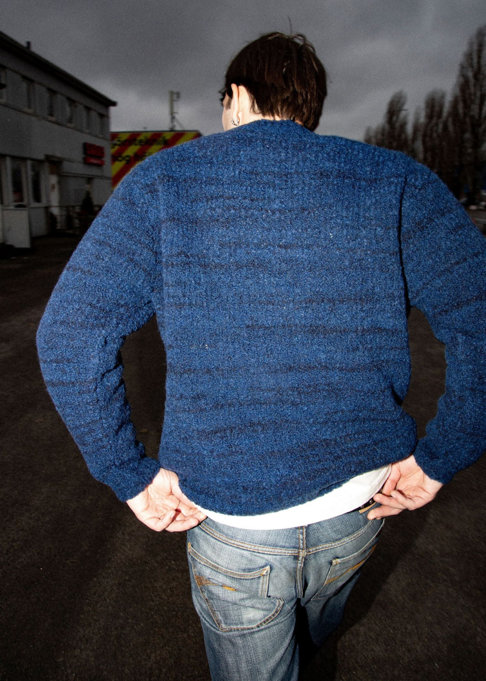 A man viewed from behind wearing a textured blue knit sweater over a white t-shirt and blue jeans, standing outdoors on a paved area with buildings and trees in the background under a cloudy sky.