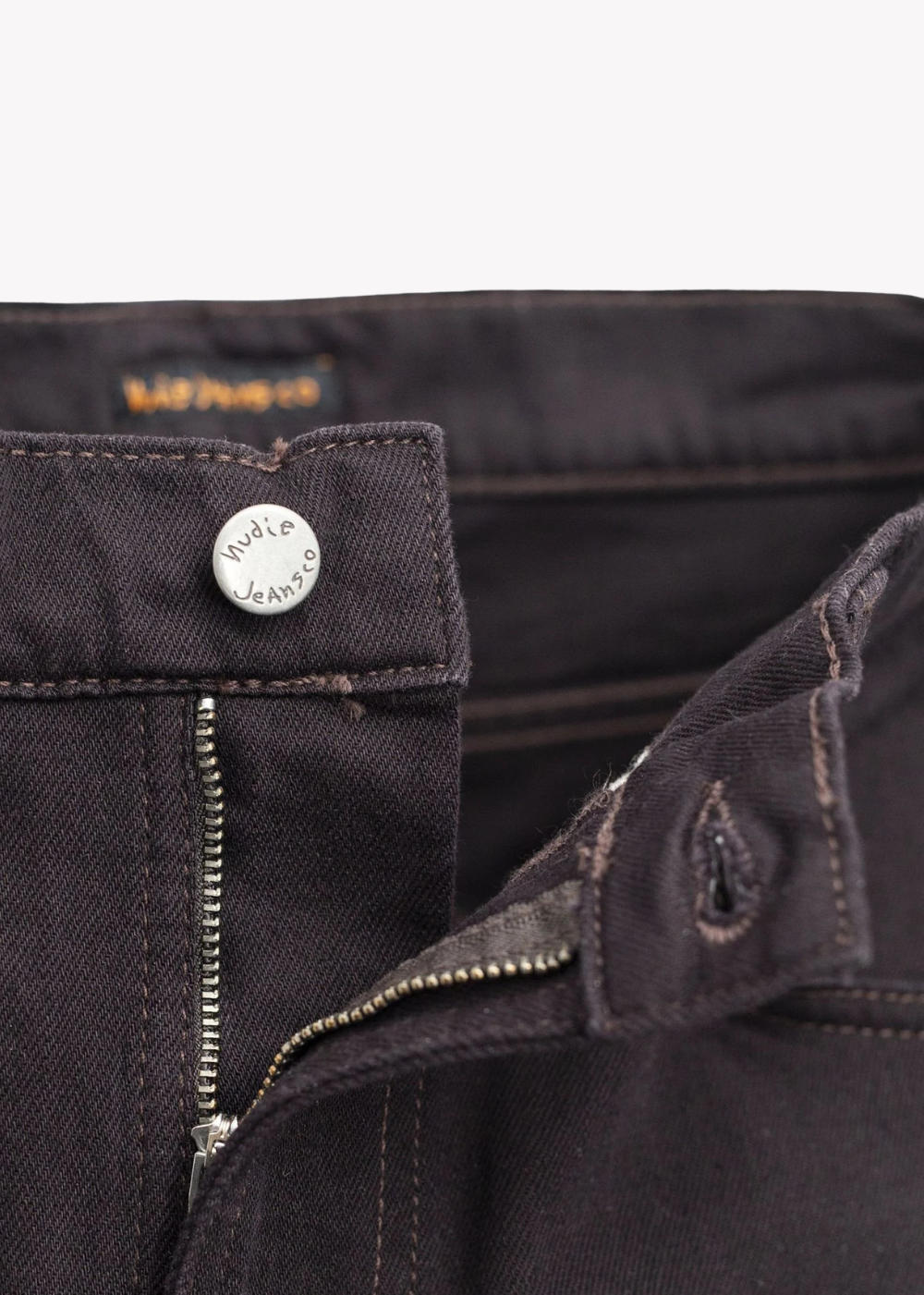 Close-up of dark brown denim jeans showing the waistband, a silver metal button with 'Waffle Jeans' branding, a zippered fly, and contrast stitching.