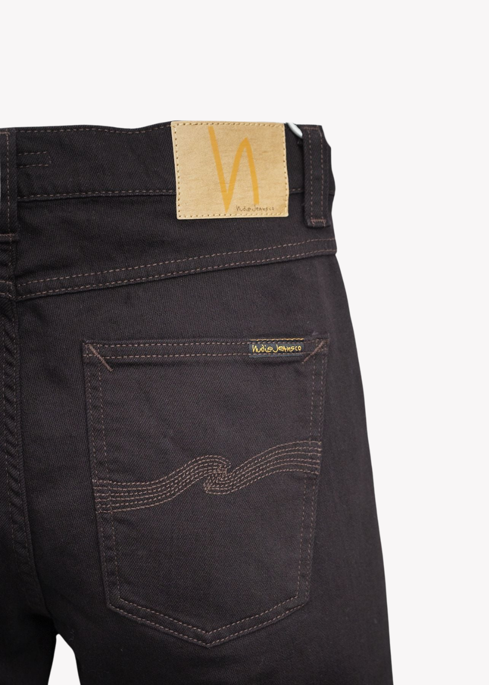 Close-up of the back waistband and pocket of dark brown denim jeans, showing a tan leather patch with a yellow 'N' logo and a small 'Willy Wonka' embroidered tag on the pocket.