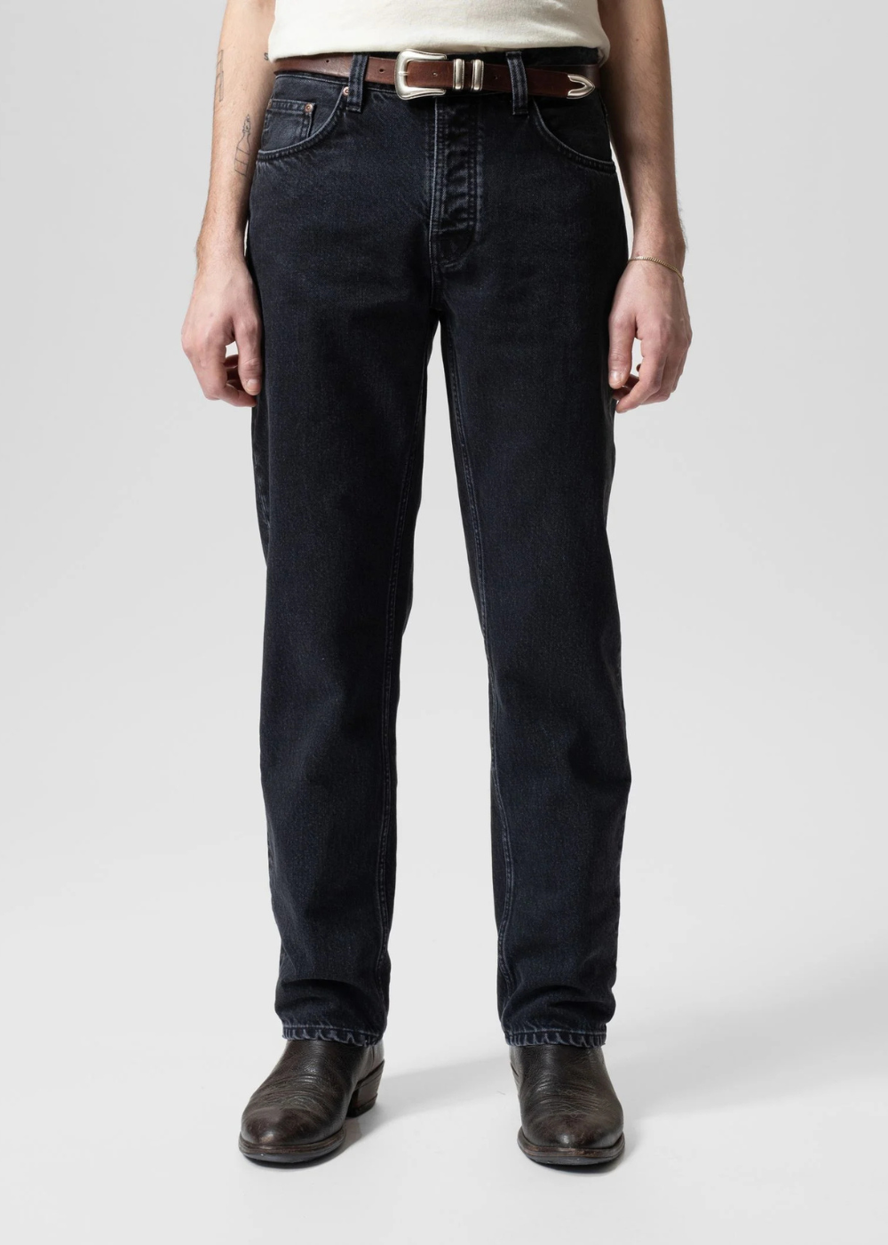 A person wearing dark indigo straight-leg jeans with a brown leather belt, paired with a cream top and dark brown leather boots, shown from the waist down on a light gray studio background.