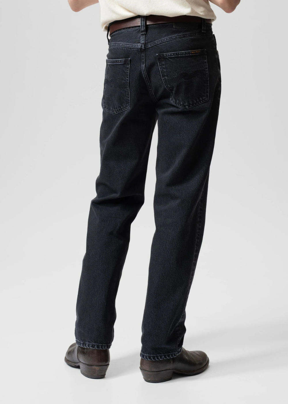 Rear view of a person wearing dark indigo straight-leg jeans with a brown belt and dark brown leather shoes, standing against a light gray studio background.