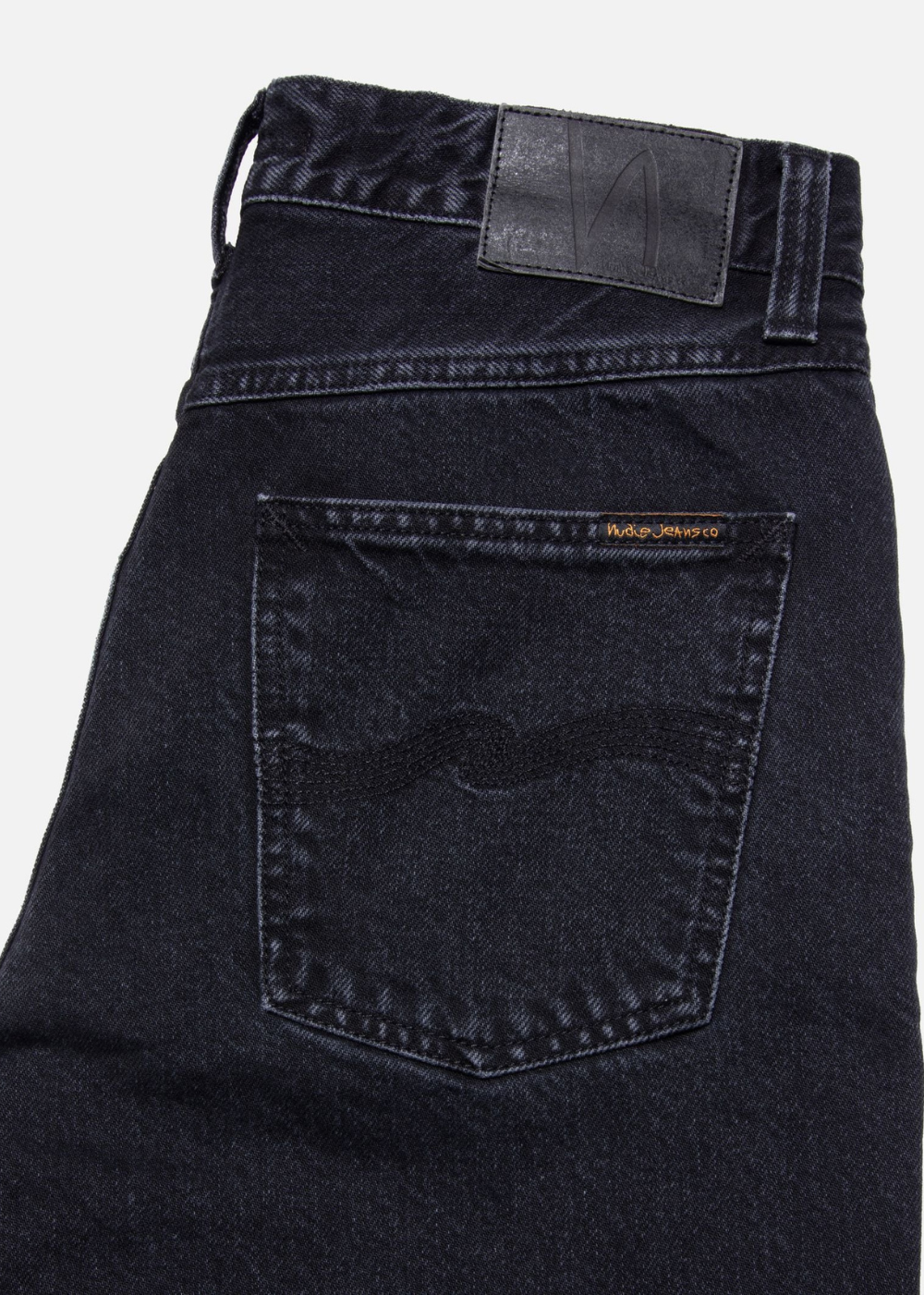Close-up of the back waistband and pocket area of dark indigo denim jeans, showing a black leather patch at the back waist and a small orange brand tab on the pocket.