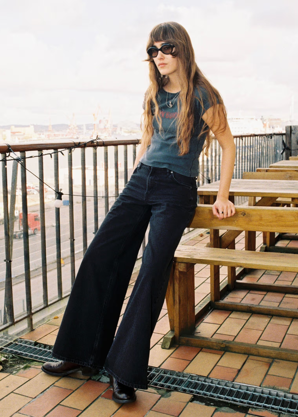 A woman wearing a faded blue graphic t-shirt, dark wide-leg jeans, and sunglasses stands on a rooftop terrace with wooden tables and a railing.