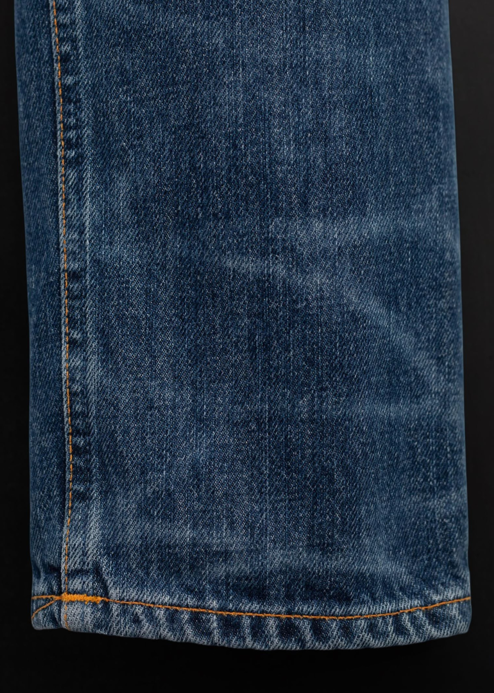 Close-up of a blue denim fabric with visible whiskering and orange contrast stitching along the hem, set against a black background.