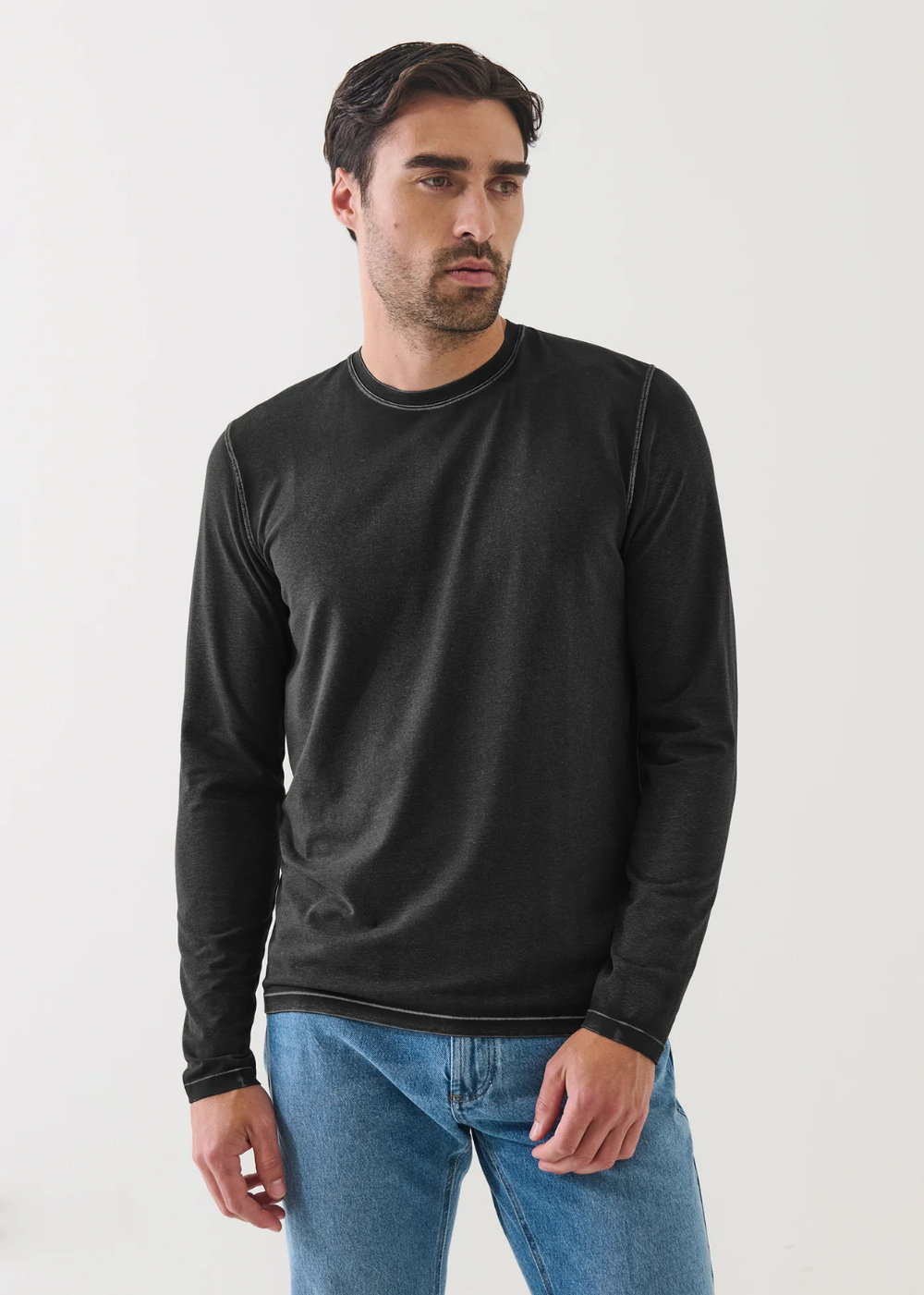 A man wearing a black long-sleeve crewneck shirt and blue jeans, standing against a plain light gray background.