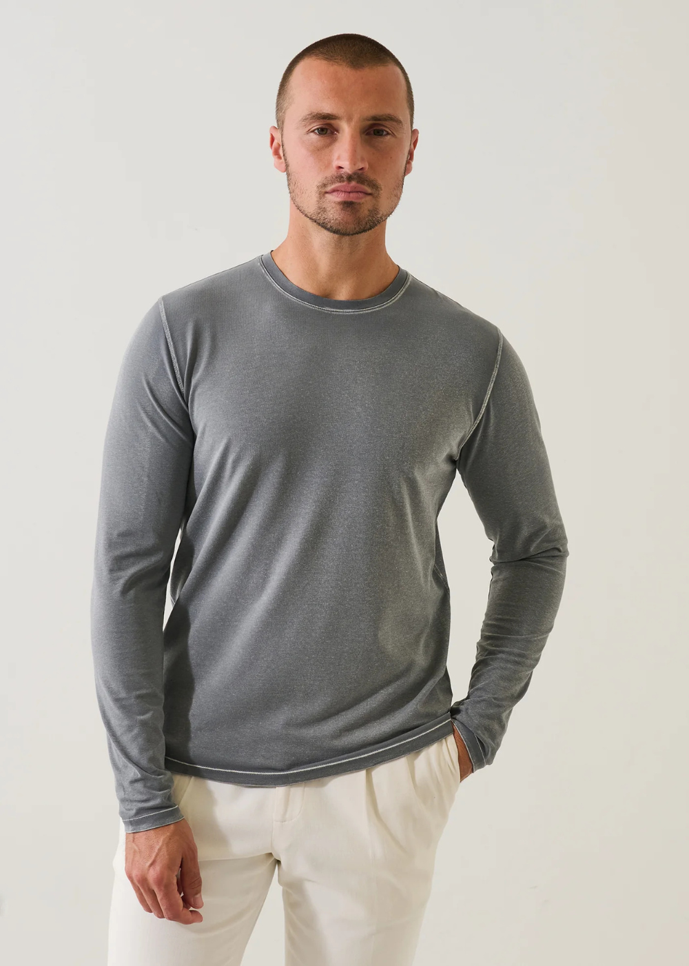 A man wearing a gray long-sleeve crewneck shirt and off-white trousers, standing against a plain light gray studio background.