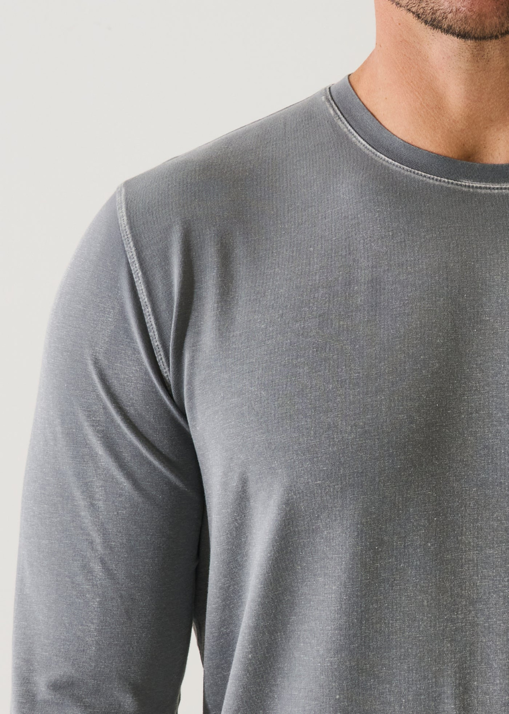 Close-up of a man wearing a heather gray long-sleeve crewneck shirt, showing the ribbed texture and collar detail against a light gray background.