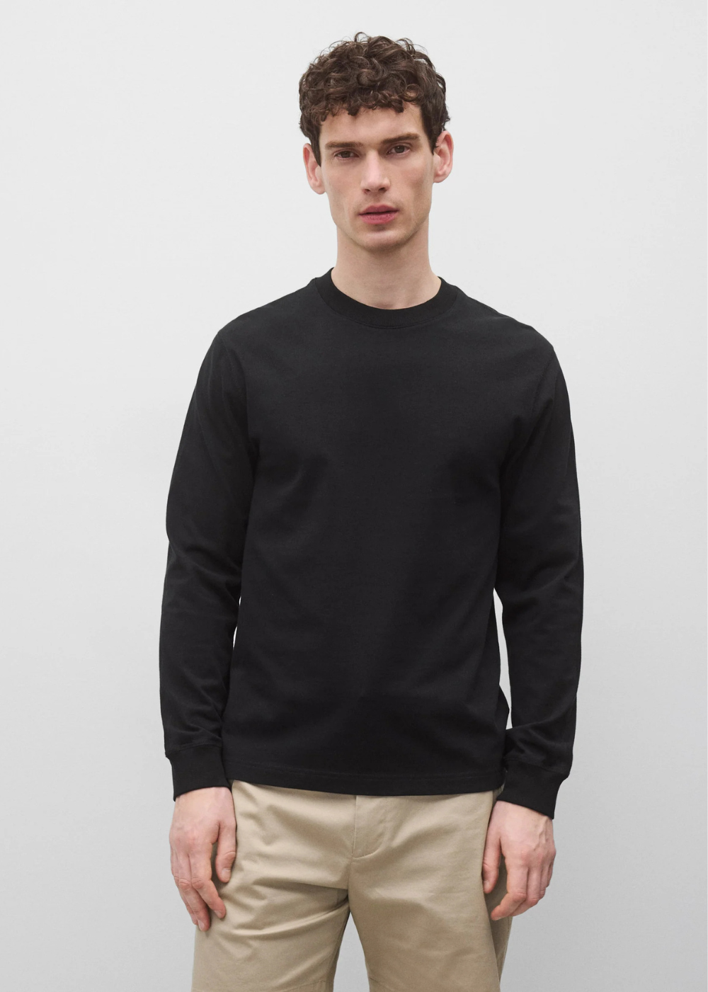 A man wearing a plain black long-sleeve crewneck sweater and beige trousers, standing against a light gray studio background.