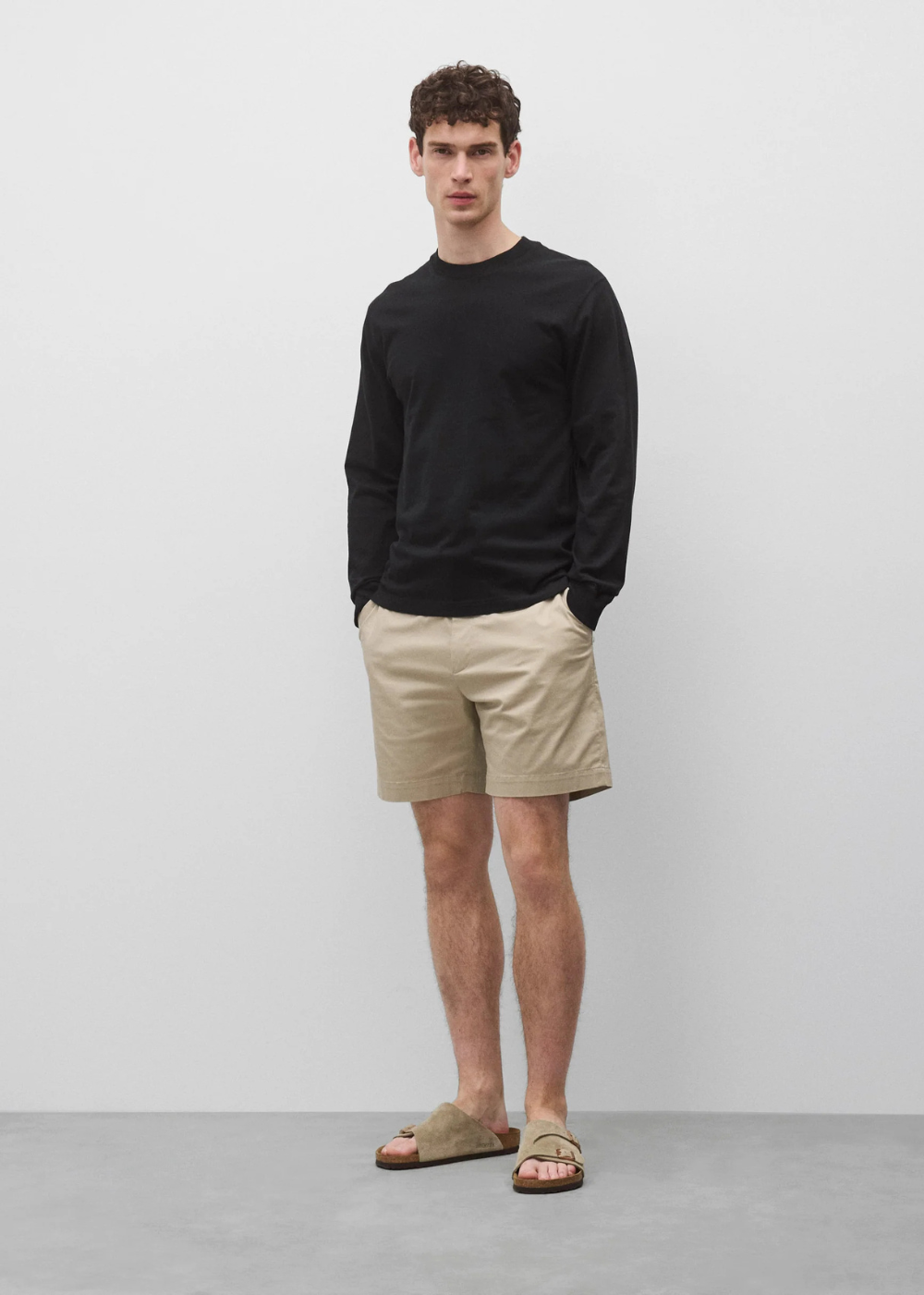 A man wearing a black long-sleeve crewneck shirt, beige shorts, and tan slide sandals, standing against a light gray studio background.