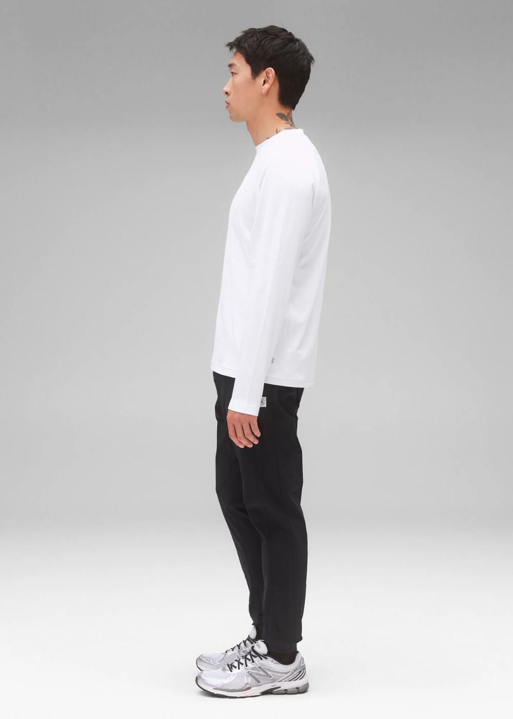 A man shown in profile wearing a white long-sleeve crewneck shirt, black pants, and silver athletic sneakers against a light gray studio background.