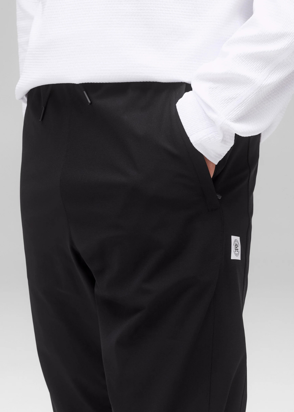 Close-up of a person wearing black trousers with a white long-sleeve shirt tucked in, showing the side pocket and a small white brand tag on the trouser leg.