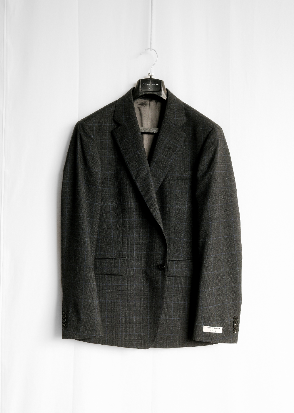 A dark charcoal gray plaid blazer jacket hanging on a black hanger against a white fabric backdrop.