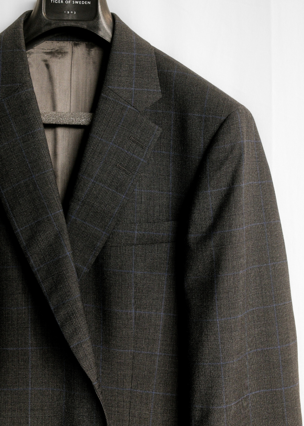 A close-up of a dark gray tweed blazer with a subtle windowpane check pattern, hanging on a black hanger labeled 'TWEED OF SWEDEN'.