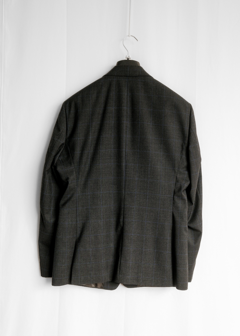 A dark charcoal gray plaid blazer jacket hanging on a black hanger against a white paneled wall, showing the back view.