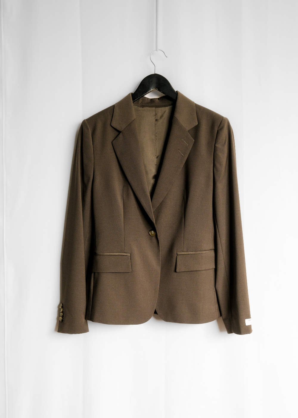 A brown single-breasted blazer jacket with notch lapels and flap pockets, hanging on a black hanger against a white fabric backdrop.