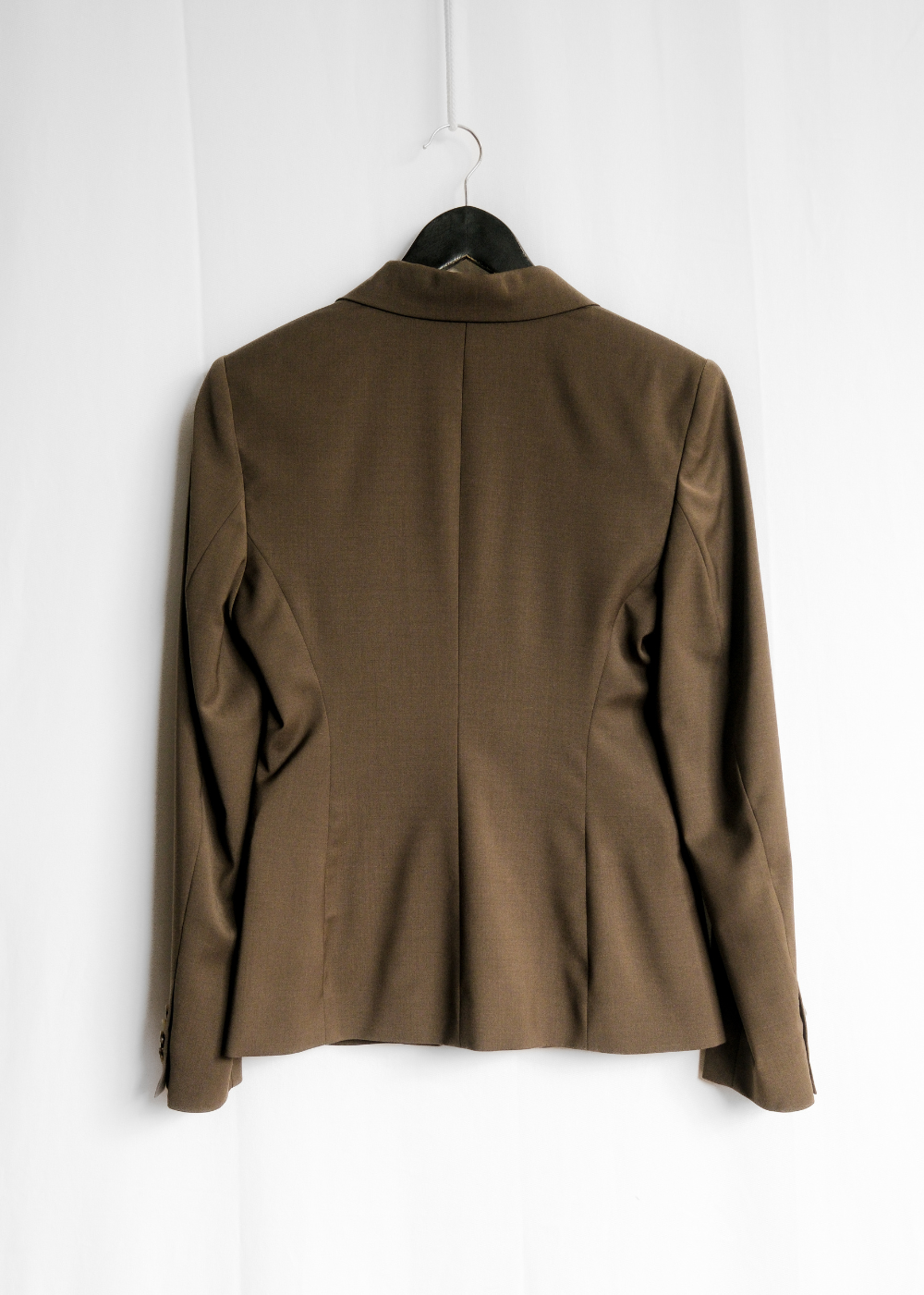 A brown tailored blazer jacket hanging on a black hanger against a white paneled wall, shown from the back.