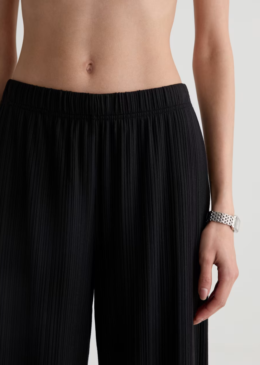Close-up of a model wearing black pleated wide-leg trousers, showing the textured fabric and waistband, with a silver watch on the wrist against a light gray background.