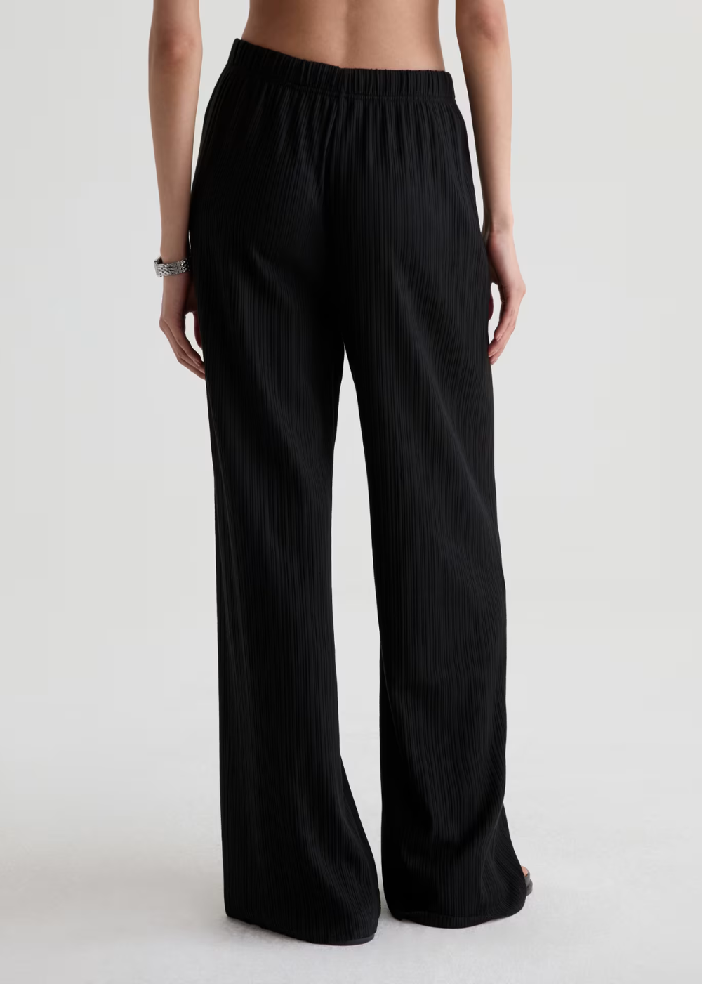Black wide-leg pleated trousers shown from the back on a model, featuring an elastic waistband and a flowing silhouette, against a plain light gray background.