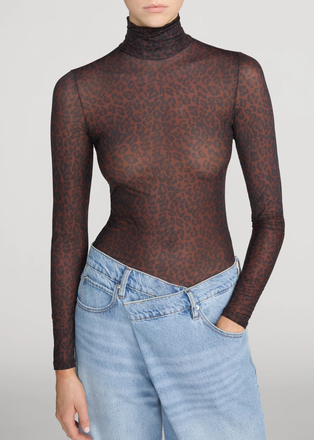 A model wearing a sheer, long-sleeve mock neck top with a dark brown and black leopard print, paired with light blue jeans, against a light gray studio background.