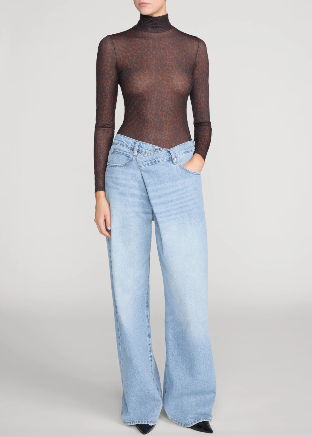A model wearing a sheer dark brown long-sleeve mock neck top and light-wash wide-leg denim jeans, standing against a neutral light gray studio background.