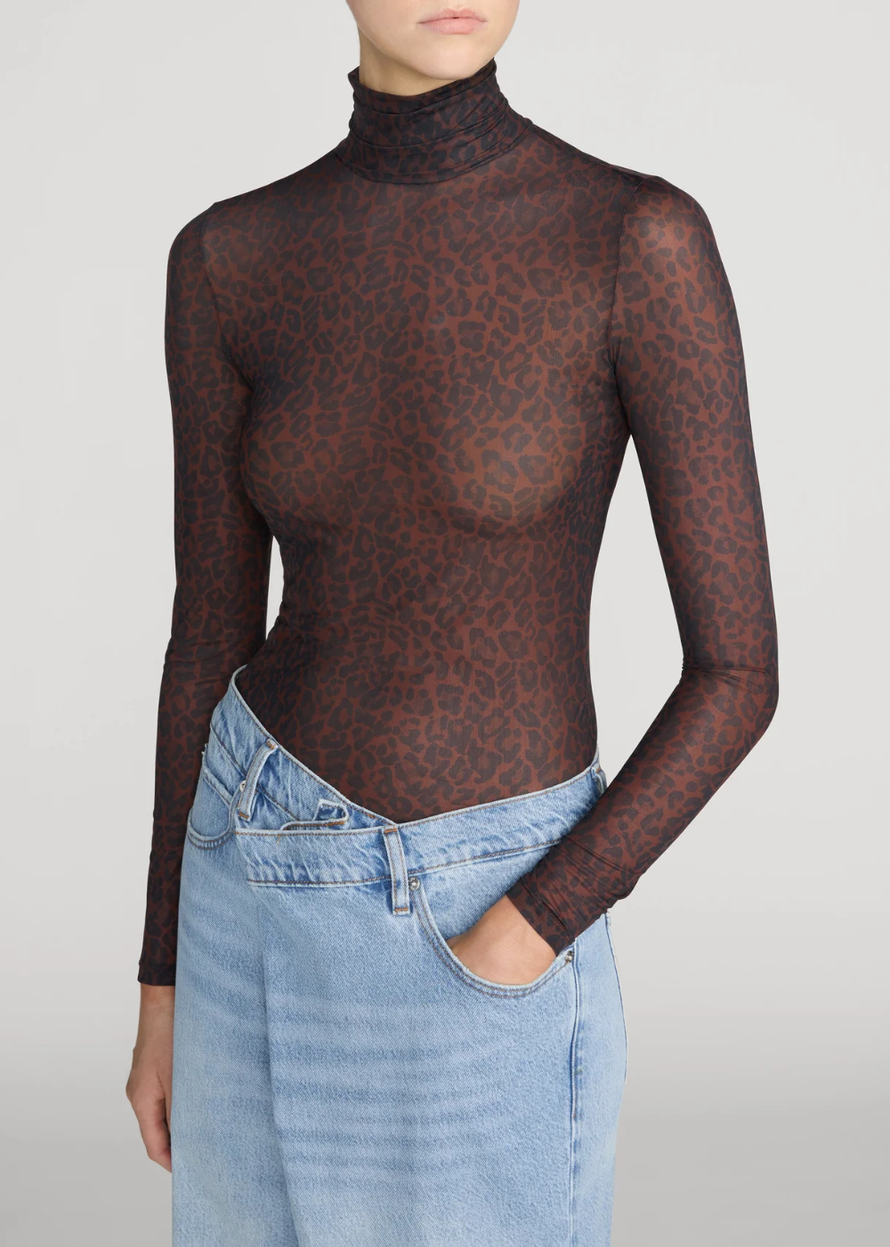 A model wearing a sheer, long-sleeve mock neck top with a dark brown and black leopard print, paired with light blue denim jeans against a light gray studio background.
