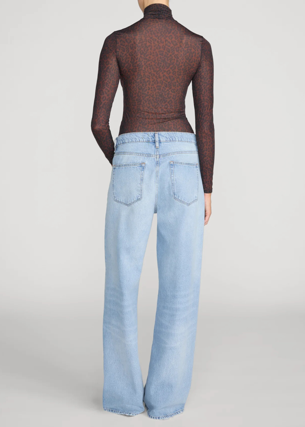 A model shown from the back wearing a sheer dark brown leopard-print long-sleeve mock neck top and light-wash wide-leg blue jeans against a light gray studio background.