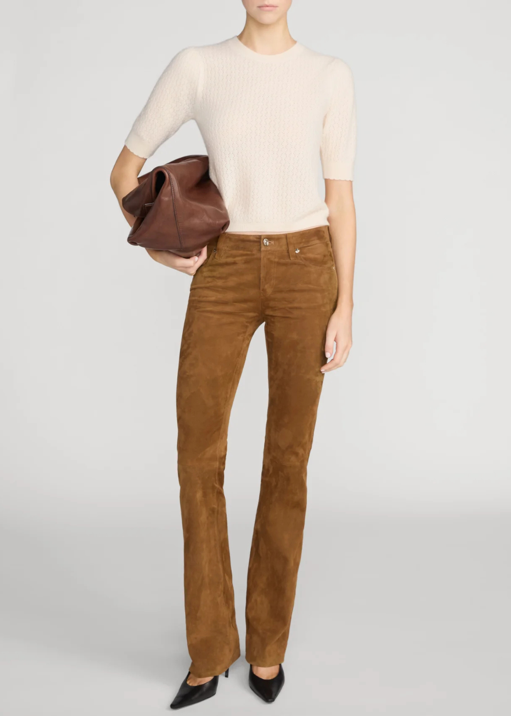 A model wearing a cream short-sleeve ribbed knit top, brown suede flare pants, and black pointed-toe heels, holding a brown leather clutch against a light gray studio background.