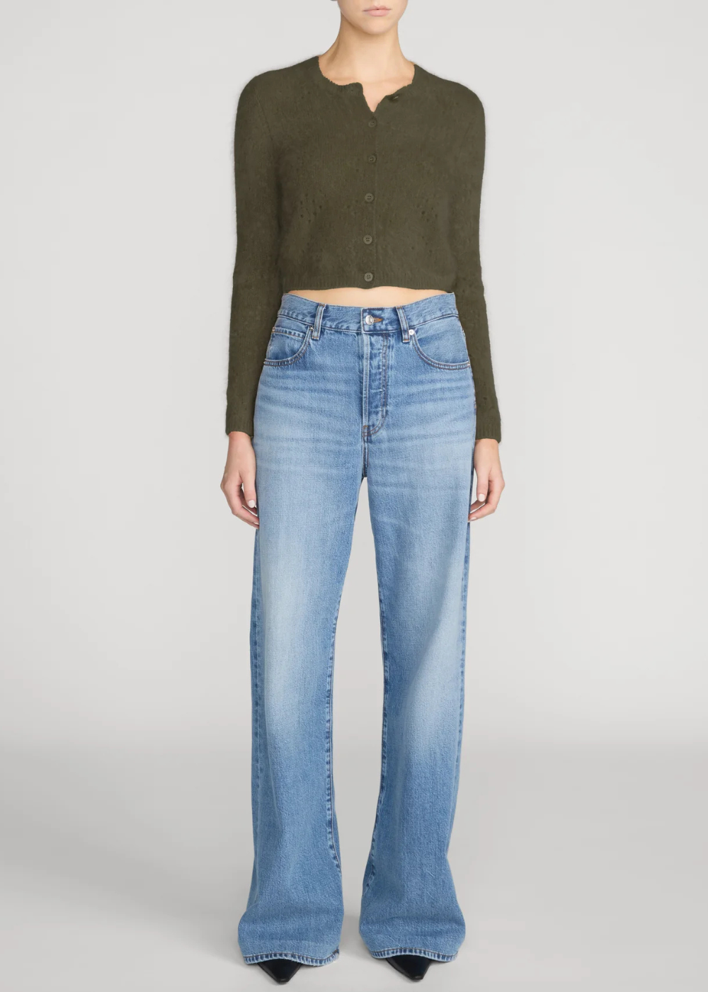 A model wearing a dark olive green cropped button-up cardigan and light-wash blue wide-leg jeans, standing against a light gray studio background.