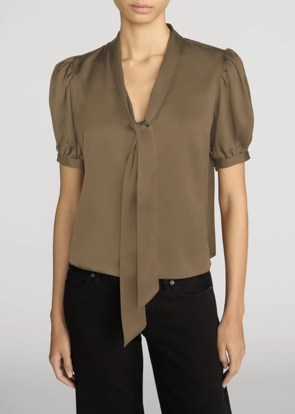 A woman wearing a taupe-brown short-sleeve blouse with puff sleeves and a front tie detail, paired with black pants, against a light gray studio background.