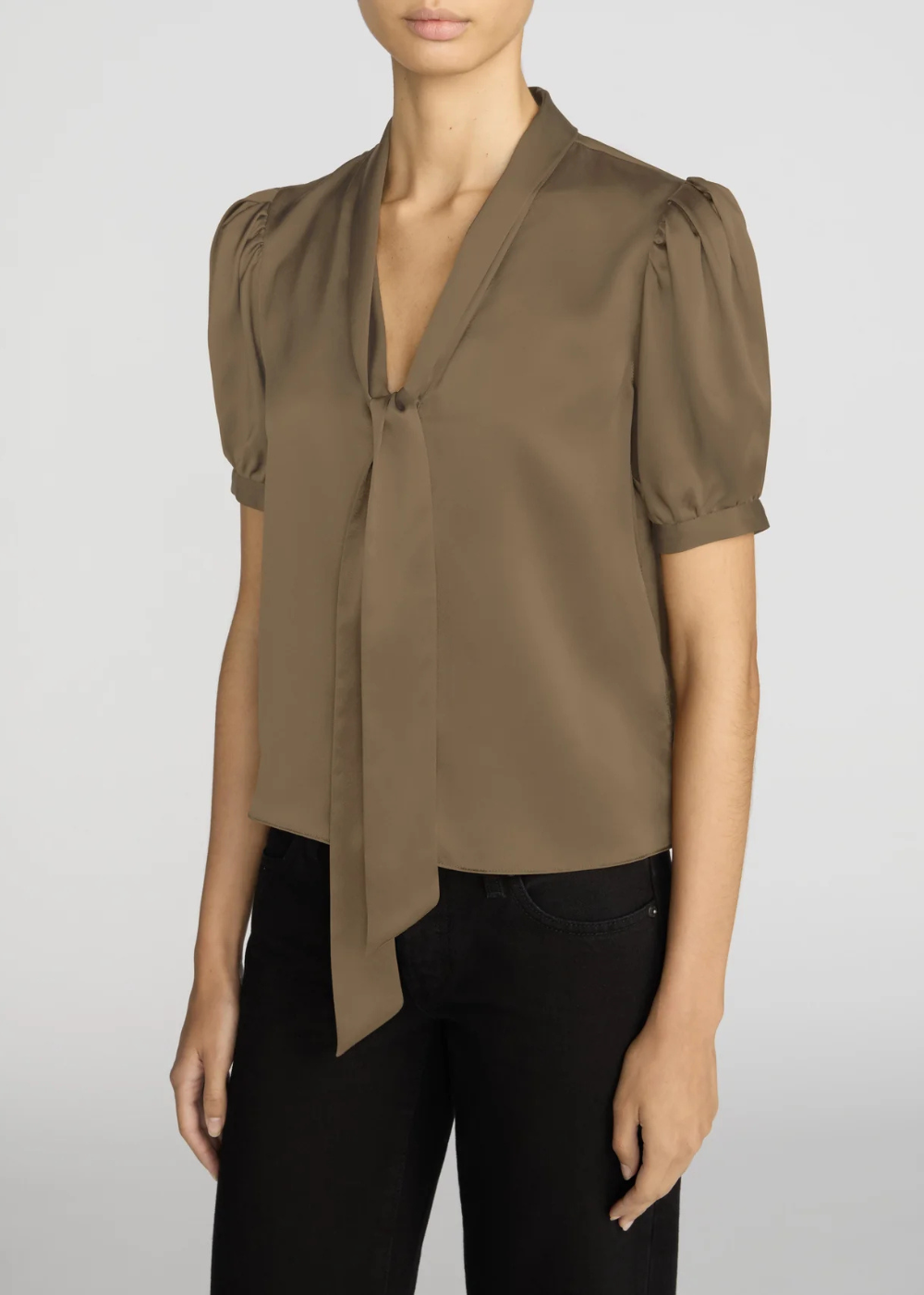 A woman wearing a taupe-brown short-sleeve blouse with puff sleeves and a front tie detail, paired with black pants, against a light gray studio background.