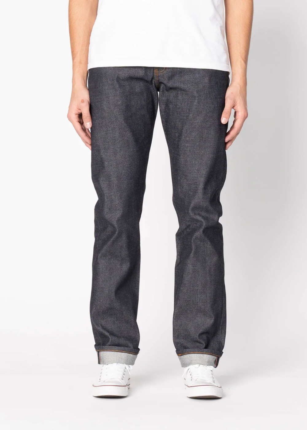 A person wearing dark indigo slim-fit jeans with a white t-shirt and white sneakers, shown from the waist down against a light gray studio background.