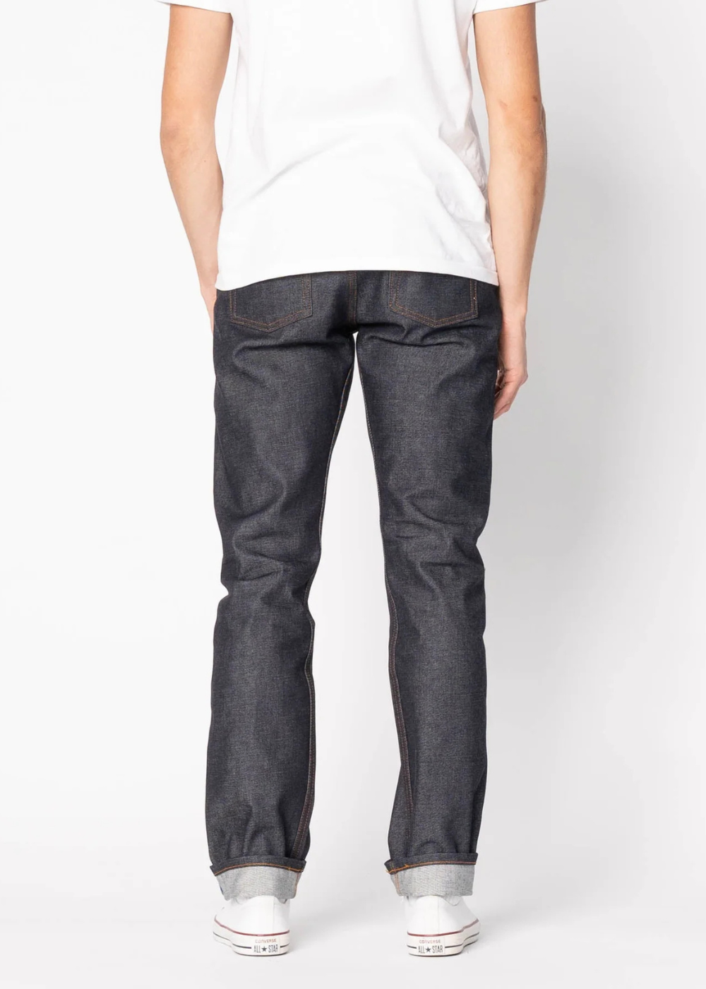 Rear view of a person wearing dark indigo slim-fit jeans, a white t-shirt, and white sneakers against a light gray studio background.
