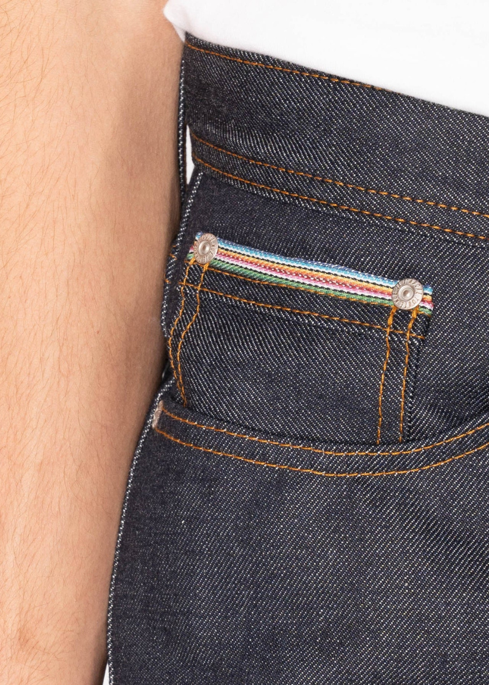 Close-up of the back pocket of dark indigo denim jeans worn by a model, showing orange contrast stitching and a colorful striped fabric tab.