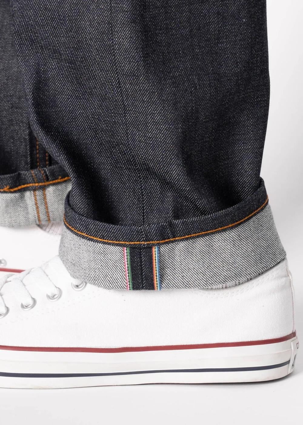 Close-up of dark indigo denim jeans rolled up at the cuff, showing the contrast stitching and hem, paired with white low-top canvas sneakers on a light gray background.